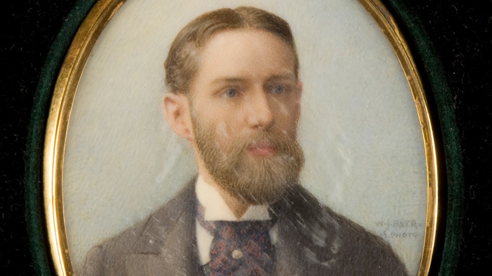 portrait in a gold oval necklace of an adult wearing a grey suit with parted hair and beard